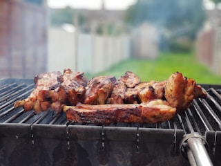 A barbecue grill with several pieces of meat being cooked, surrounded by a smoky atmosphere. The background shows a blurred outdoor setting with grass and a fence.