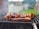 A barbecue grill with several pieces of meat being cooked, surrounded by a smoky atmosphere. The background shows a blurred outdoor setting with grass and a fence.