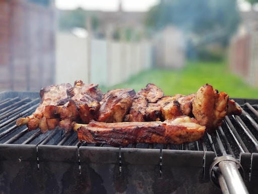 A barbecue grill with several pieces of meat being cooked, surrounded by a smoky atmosphere. The background shows a blurred outdoor setting with grass and a fence.