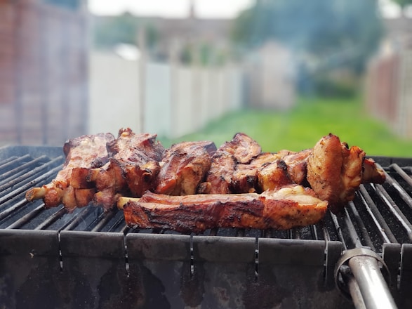 A barbecue grill with several pieces of meat being cooked, surrounded by a smoky atmosphere. The background shows a blurred outdoor setting with grass and a fence.