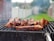 A barbecue grill with several pieces of meat being cooked, surrounded by a smoky atmosphere. The background shows a blurred outdoor setting with grass and a fence.