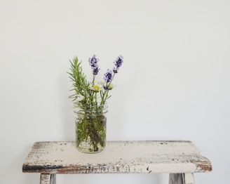 A small glass jar filled with sprigs of lavender, rosemary, and a few white daisies is placed on a rustic wooden plank with a faded, distressed white paint finish. The background is a plain, light-colored wall, creating a simple and minimalist composition.