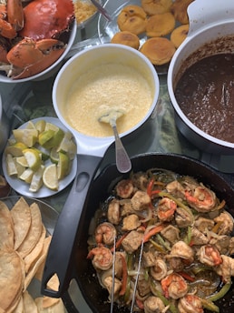 A table spread with assorted dishes including a bowl of cooked crab, a skillet filled with shrimp, chicken, and sliced vegetables, a plate of fried plantains, and bowls of sauces. Lime wedges and tortilla chips are also visible.