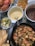 A table spread with assorted dishes including a bowl of cooked crab, a skillet filled with shrimp, chicken, and sliced vegetables, a plate of fried plantains, and bowls of sauces. Lime wedges and tortilla chips are also visible.