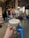 A hand holds a cocktail glass filled with a frothy, light-colored drink. Behind it, the interior of a distillery is visible, featuring copper distillation equipment and various containers. There is a blue stool and a forklift in the background, giving an industrial yet cozy ambience.