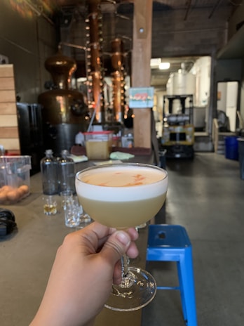 A hand holds a cocktail glass filled with a frothy, light-colored drink. Behind it, the interior of a distillery is visible, featuring copper distillation equipment and various containers. There is a blue stool and a forklift in the background, giving an industrial yet cozy ambience.