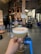 A hand holds a cocktail glass filled with a frothy, light-colored drink. Behind it, the interior of a distillery is visible, featuring copper distillation equipment and various containers. There is a blue stool and a forklift in the background, giving an industrial yet cozy ambience.