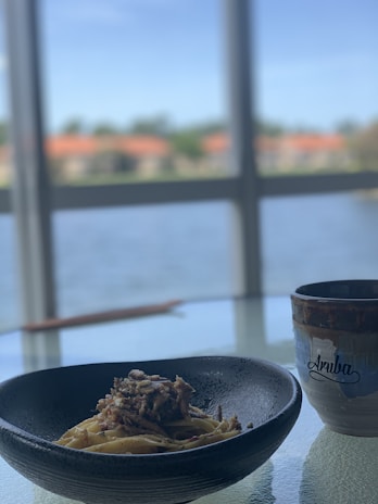 A bowl of pasta topped with a savory mixture sits on a glass table next to a cup with 'Aruba' written on it. Through the windows, a blurred view of water and buildings with red roofs can be seen under a clear blue sky.