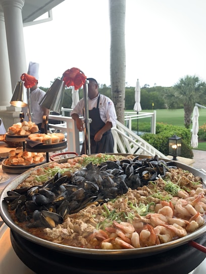 A large outdoor food display featuring a paella pan filled with a variety of fresh seafood, including mussels, shrimp, and pieces of fish, garnished with green onions. Behind the paella are stacks of bread rolls and two chefs in white uniforms preparing more food items. The setting appears to be a terrace overlooking a green lawn with palm trees, creating a tropical atmosphere.
