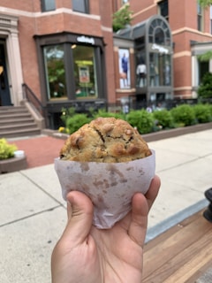 A hand holding a large, freshly baked chocolate chip cookie wrapped in a piece of white parchment paper. The background shows an urban street scene with red brick buildings, stairs, and a storefront with large windows. Green shrubbery and planters line the sidewalk, adding a touch of nature to the city setting.