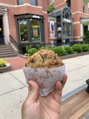 A hand holding a large, freshly baked chocolate chip cookie wrapped in a piece of white parchment paper. The background shows an urban street scene with red brick buildings, stairs, and a storefront with large windows. Green shrubbery and planters line the sidewalk, adding a touch of nature to the city setting.