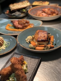 A selection of gourmet dishes is artfully presented on a table. The main focus is a glazed fish or meat dish with carrots and cauliflower, served on a blue plate. Surrounding it are various other dishes, including what appears to be fried chicken on a dark rectangular plate with a side salad, and other entrees elegantly plated. The lighting highlights the textures and colors of the dishes, enhancing their appetizing appearance.