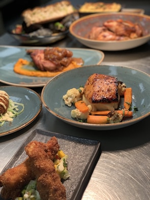 A selection of gourmet dishes is artfully presented on a table. The main focus is a glazed fish or meat dish with carrots and cauliflower, served on a blue plate. Surrounding it are various other dishes, including what appears to be fried chicken on a dark rectangular plate with a side salad, and other entrees elegantly plated. The lighting highlights the textures and colors of the dishes, enhancing their appetizing appearance.