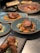 A selection of gourmet dishes is artfully presented on a table. The main focus is a glazed fish or meat dish with carrots and cauliflower, served on a blue plate. Surrounding it are various other dishes, including what appears to be fried chicken on a dark rectangular plate with a side salad, and other entrees elegantly plated. The lighting highlights the textures and colors of the dishes, enhancing their appetizing appearance.