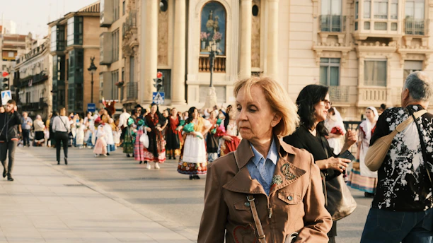A group of people, some in traditional and colorful attire, are gathered in a public square near historical buildings. A woman in a brown jacket walks in the foreground, while other individuals, including photographers, can be seen in the background.