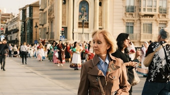 A group of people, some in traditional and colorful attire, are gathered in a public square near historical buildings. A woman in a brown jacket walks in the foreground, while other individuals, including photographers, can be seen in the background.