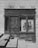 An old wooden storefront with weathered doors and windows is set against a stone wall. A poster in the window advertises live jazz music with a visible saxophone image and text. Stone steps with a shadow leading up to the door create a sense of age and history.