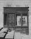 An old wooden storefront with weathered doors and windows is set against a stone wall. A poster in the window advertises live jazz music with a visible saxophone image and text. Stone steps with a shadow leading up to the door create a sense of age and history.