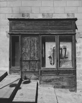 An old wooden storefront with weathered doors and windows is set against a stone wall. A poster in the window advertises live jazz music with a visible saxophone image and text. Stone steps with a shadow leading up to the door create a sense of age and history.