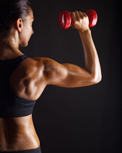 A person with well-defined muscles is lifting a red dumbbell, showcasing strength and fitness against a dark background. The individual's skin is glistening, highlighting their muscular build.