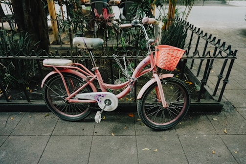 A pink children's bicycle with a basket is parked on a sidewalk next to an iron fence and greenery. The bike is secured with a chain, and there are fallen leaves scattered on the pavement nearby.