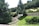 A scenic outdoor landscape featuring a variety of green trees and shrubs. A large tree stands prominently in the center, surrounded by well-manicured bushes and grass. Stone steps lead upwards, flanked by stone walls, creating a structured garden layout. The background is filled with a dense forest of evergreens.