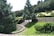 A scenic outdoor landscape featuring a variety of green trees and shrubs. A large tree stands prominently in the center, surrounded by well-manicured bushes and grass. Stone steps lead upwards, flanked by stone walls, creating a structured garden layout. The background is filled with a dense forest of evergreens.