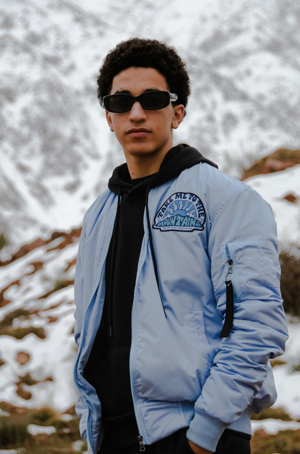 a man in a blue jacket and sunglasses standing in front of a mountain