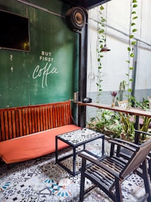A cozy outdoor seating area with a green-painted wall featuring a quote about coffee, a wooden bench with a bright orange cushion, a patterned tiled floor, and a black metal-framed table with a matching chair. Potted plants with trailing vines and a hanging lamp add a touch of greenery and warmth.