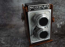 A vintage twin-lens reflex camera with metal dials and a worn leather covering. The camera is marked with 'Ricohflex Model VI' and features two large lenses on the front, surrounded by engraved text indicating specifications. The background is a textured, dark surface, which contrasts with the metallic elements of the camera.