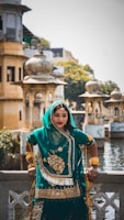 A woman in traditional Indian attire is standing by a decorative railing with intricate architectural structures in the background. She is wearing a teal and gold embellished outfit with matching jewelry. The setting appears to be a historical or culturally significant site with domed structures and a water body nearby.