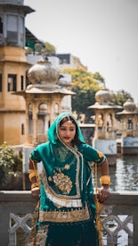A woman in traditional Indian attire is standing by a decorative railing with intricate architectural structures in the background. She is wearing a teal and gold embellished outfit with matching jewelry. The setting appears to be a historical or culturally significant site with domed structures and a water body nearby.