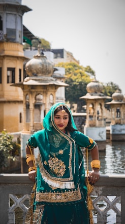 A woman in traditional Indian attire is standing by a decorative railing with intricate architectural structures in the background. She is wearing a teal and gold embellished outfit with matching jewelry. The setting appears to be a historical or culturally significant site with domed structures and a water body nearby.