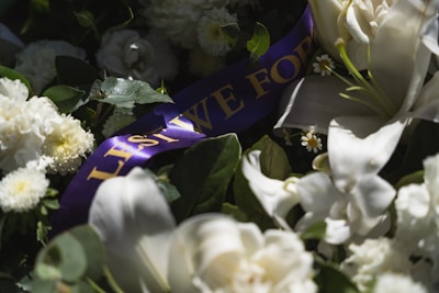 A collection of white flowers including lilies and chrysanthemums is arranged with green foliage. Among the flowers, a piece of purple ribbon is visible with gold text partially reading 'LEST WE FOR...'. The overall composition is illuminated by soft, natural light.