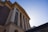 A building with large classical columns and a banner reading 'Faculty of Medicine' hanging between two columns. The structure has a mix of brick and stonework with an intricate cornice at the top. The sky is clear with a bright sun illuminating the scene from behind the building.