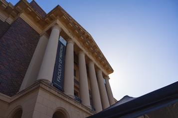 A building with large classical columns and a banner reading 'Faculty of Medicine' hanging between two columns. The structure has a mix of brick and stonework with an intricate cornice at the top. The sky is clear with a bright sun illuminating the scene from behind the building.