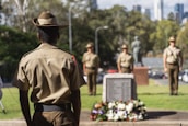 Several soldiers are standing in a formal formation near a memorial adorned with a wreath of flowers. The soldiers are wearing uniforms and hats, and they are positioned in a park-like setting with trees and a cityscape in the background.