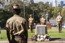 Several soldiers are standing in a formal formation near a memorial adorned with a wreath of flowers. The soldiers are wearing uniforms and hats, and they are positioned in a park-like setting with trees and a cityscape in the background.