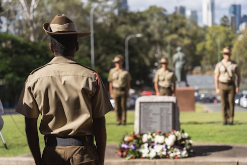 Several soldiers are standing in a formal formation near a memorial adorned with a wreath of flowers. The soldiers are wearing uniforms and hats, and they are positioned in a park-like setting with trees and a cityscape in the background.