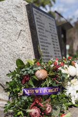 A memorial wreath adorned with green foliage and various flowers, including red and pink proteas, is placed by a stone memorial. The wreath features a purple ribbon with 'LEST WE FORGET' in gold lettering. The stone has engraved names, indicating a tribute to fallen soldiers. In the background, there are additional floral arrangements and blurred greenery.