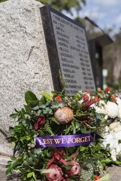 A memorial wreath adorned with green foliage and various flowers, including red and pink proteas, is placed by a stone memorial. The wreath features a purple ribbon with 'LEST WE FORGET' in gold lettering. The stone has engraved names, indicating a tribute to fallen soldiers. In the background, there are additional floral arrangements and blurred greenery.
