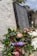 A memorial wreath adorned with green foliage and various flowers, including red and pink proteas, is placed by a stone memorial. The wreath features a purple ribbon with 'LEST WE FORGET' in gold lettering. The stone has engraved names, indicating a tribute to fallen soldiers. In the background, there are additional floral arrangements and blurred greenery.
