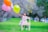 A young child wearing a floral dress stands on a small wooden chair in a grassy park, joyfully reaching for a cluster of colorful balloons. The background is a blur of trees and a pathway, suggesting a peaceful outdoor setting.