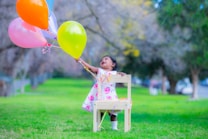 A young child wearing a floral dress stands on a small wooden chair in a grassy park, joyfully reaching for a cluster of colorful balloons. The background is a blur of trees and a pathway, suggesting a peaceful outdoor setting.