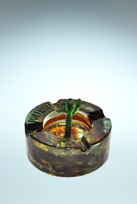 A glass ashtray with an amber and green color pattern featuring a small cactus-shaped sculptural element in the center. The ashtray has a textured and glossy surface, adding a vintage or decorative appeal.