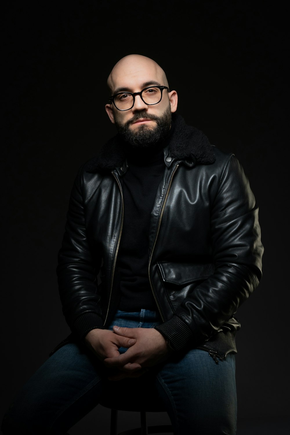 a man in a black jacket and glasses sitting on a stool