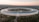 A large, circular building with a futuristic design is surrounded by greenery and trees. The structure features extensive solar panels on its roof. There is a spacious landscaped area in the middle, and the horizon shows a blend of natural light from the setting or rising sun.