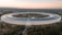 A large, circular building with a futuristic design is surrounded by greenery and trees. The structure features extensive solar panels on its roof. There is a spacious landscaped area in the middle, and the horizon shows a blend of natural light from the setting or rising sun.