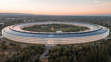 A large, circular building with a futuristic design is surrounded by greenery and trees. The structure features extensive solar panels on its roof. There is a spacious landscaped area in the middle, and the horizon shows a blend of natural light from the setting or rising sun.