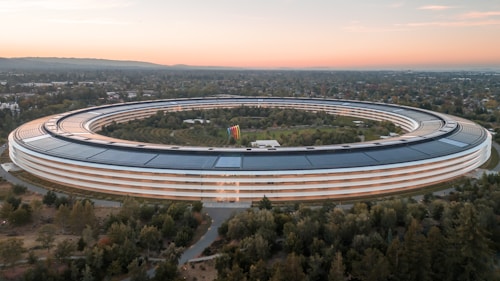 A large, circular building with a futuristic design is surrounded by greenery and trees. The structure features extensive solar panels on its roof. There is a spacious landscaped area in the middle, and the horizon shows a blend of natural light from the setting or rising sun.